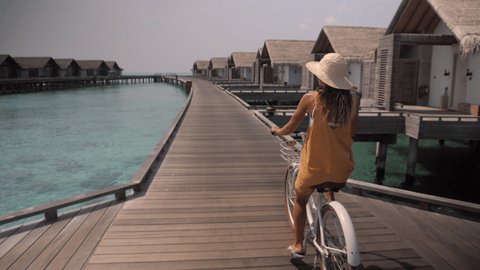 Young woman riding bicycle around tourist luxury resort in the Maldive Island. Overwater bungalows, female enjoys tropical vacation 