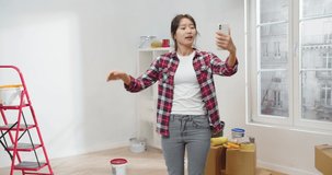Beautiful happy young Asian female videochatting online on smartphone during home renovation standing in room with red ladder and many boxes. Moving in new apartment, repair and makeover
