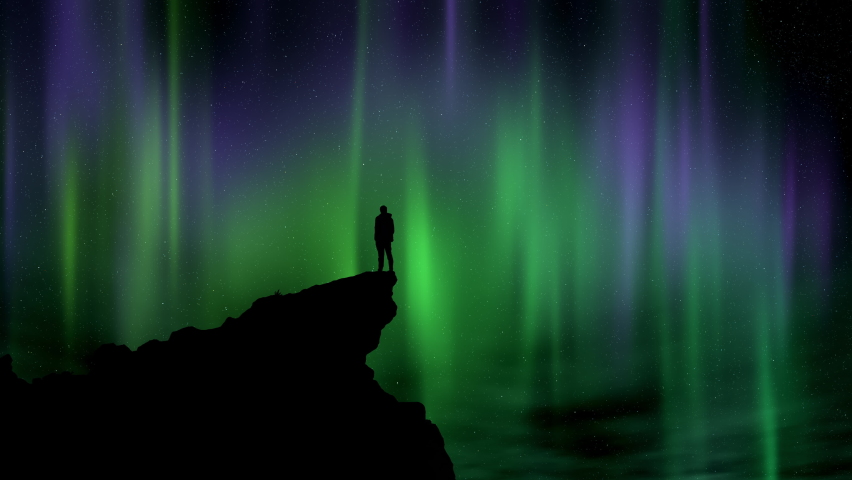 The silhouette of a man standing on a cliff edge watching the night sky and Aurora Borealis.