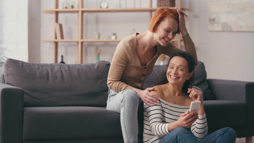 Multicultural lesbian couple looking at smartphone and smiling in living room | Shutterstock HD Video #1067714879