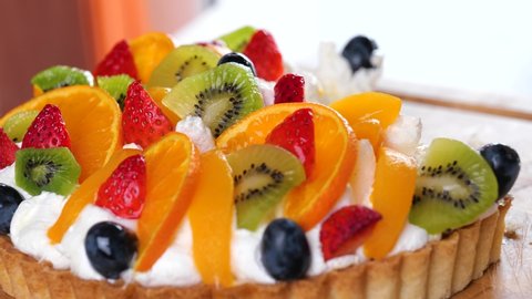 woman hand cuts with knife a delicious beautiful fruit cake with whipped cream, kiwi, grape, apricot, strawberries, orange slices. Dessert, sweet delicious holiday treats. Slow motion