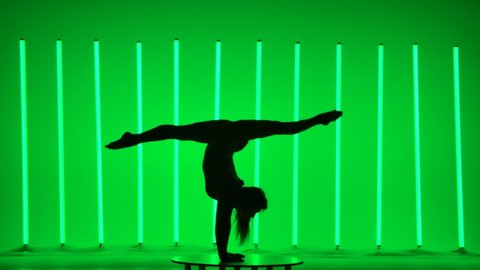 Side view of a dark silhouette of a young woman gymnast in the studio against a background of bright multicolored neon lights. Girl does a handstand in slow motion.