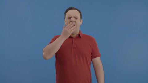 IThe young man is yawning, drowsy. Yawning young caucasian man, showing a tired gesture, covering the mouth with his hand. ndoor studio shot isolated on blue background. 
