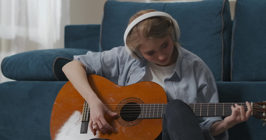 play guitar alone at home, young woman is doing exercises on guitar, listening to melody by headphones