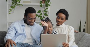 Three people parents with small daughter Afro American couple and cute preschool girl sitting on couch at home using laptop for remote communication video chat conferences online waving hands greeting