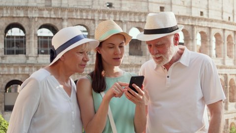 Tourists family in hats enjoy vacation in Rome. Senior couple and young teenage daughter study city map app on cellphone near famous Italian attraction Colosseum. Lost in city look for right direction