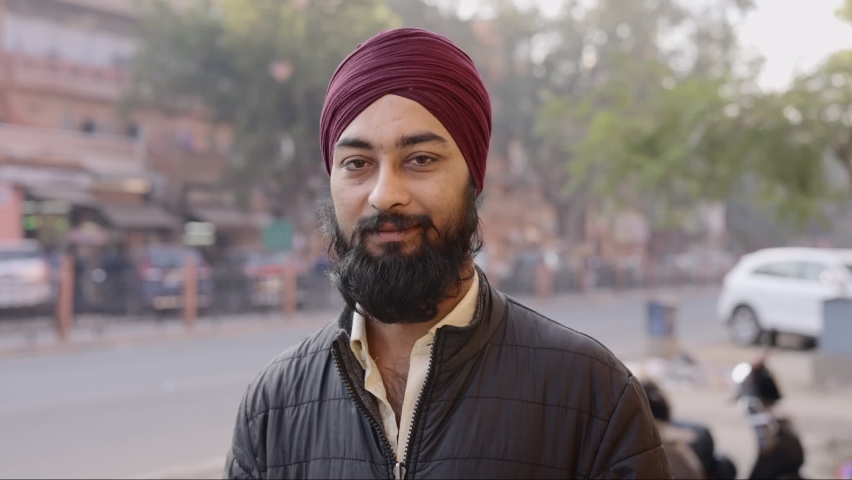 Static shot of a young Indian office worker man wearing a traditional turban on the head with a long beard looking at the camera, standing by the corner of a street during morning hours