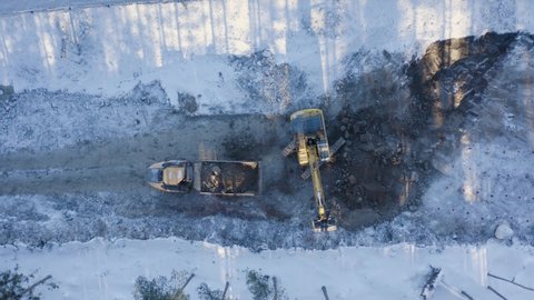 Top-down aerial view of the excavator loading soil on a dump truck in winter