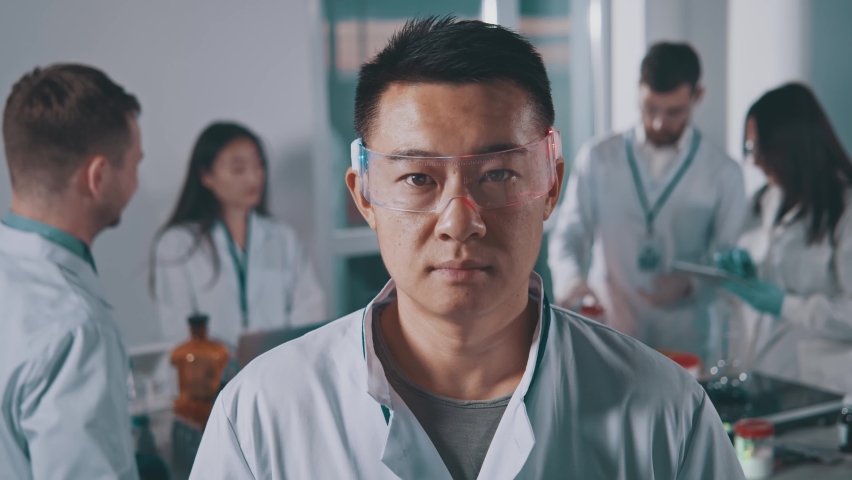 Serious young asian scientist man look at camera in protective glasses. Background team work in the laboratory. Pharmacy, microbiology, industry, biology, technology, doctor. Slow motion Royalty-Free Stock Footage #1067749151