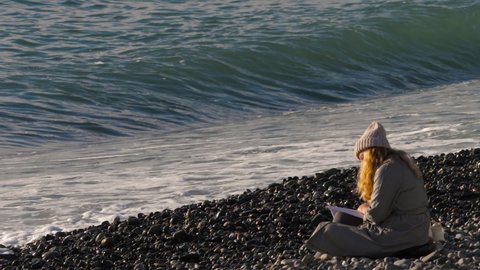 A girl in a coat and a hat sits on the seashore and reads a book