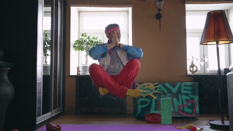 An 80s freak guy in a stylish bright tracksuit levitates over a yoga mat, doing fitness alone at home in isolation. The strange guy shows magic.