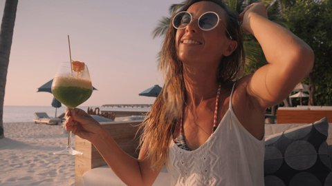 Slow motion: Woman having a cocktail at sunset on tropical beach. Young female partying in luxury establishment on the beach 