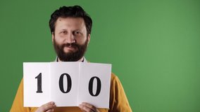 Man holding number 100 sign. Cheerful white bearded good-looking man in yellow sweater shows number one hundred on white sheet. Chrome key, green screen, copy space - Powered by Shutterstock - Get 15% off with code: PIKWIZARD15