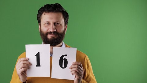 Man holding number 16 sign. Cheerful white bearded good-looking man in yellow sweater shows number sixteen on white sheet. Chrome key, green screen, copy space