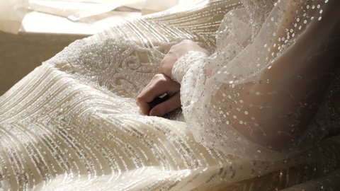 Wedding dress shiny skirt decorated with pearls and transparent sleeves with paillettes on bride at window closeup slow motion