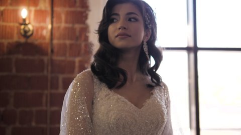 Elegant woman with earrings in sparkling wedding dress decorated with paillettes near window in restaurant closeup slow motion