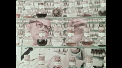1960s: Scientist grabs bottle from wall. He writes onto paper. He lifts up a beaker and smells the rods that come out of it, grabs another bottle and smells it.