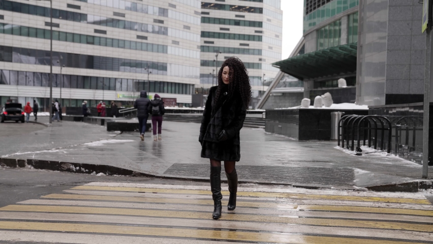A woman in a black fur coat and high-heeled boots strolls through the winter city, enjoying a beautiful day. A lady walks through a pedestrian crosswalk against the backdrop of modern buildings.