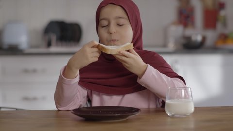 Muslim girl in a turban a llittle biting and fondly eating a cream cheese spread. Muslim girl child. Cheese bread eating concept.