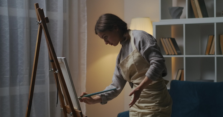 female artist is painting on canvas at home at evening, drawing picture in apartment, doing hobby at home at weekend