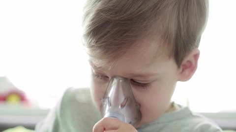 close up Caucasian 3-4 years minor preschool boy kid in the mask of an inhaler on yellow gray sofa. Baby taking respiratory therapy with nebulizer. medicine and health, pediatrics, covid-19 concept