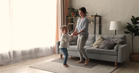 Energetic young nanny or mommy holding hands with small kid girl, jumping twisting dancing to disco music barefoot on floor carpet in living room, having fun together at home, enjoying active weekend.
