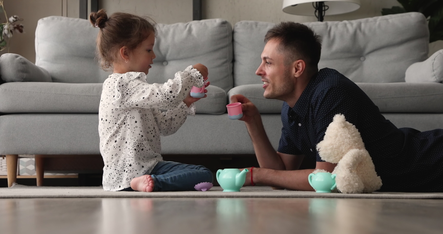 Happy small adorable preschool baby girl sitting on floor carpet, playing tea ceremony game with affectionate young father, imagining drinking from toy cups together in living room, feeling playful. Royalty-Free Stock Footage #1067779199