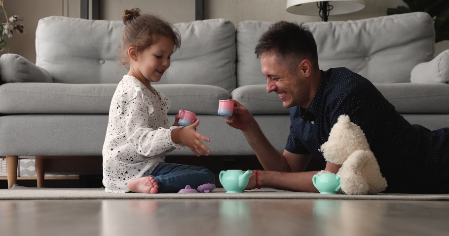 Happy small adorable preschool baby girl sitting on floor carpet, playing tea ceremony game with affectionate young father, imagining drinking from toy cups together in living room, feeling playful. | Shutterstock HD Video #1067779199