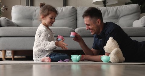 Happy small adorable preschool baby girl sitting on floor carpet, playing tea ceremony game with affectionate young father, imagining drinking from toy cups together in living room, feeling playful.