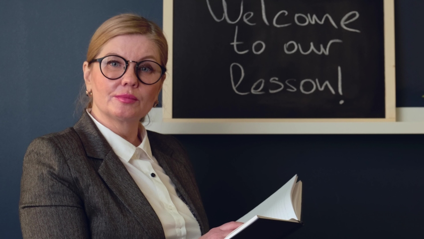 Caucasian tutor is greeting students in front of the class to her first lesson holding a book and wearing glasses