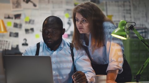 African man with holsters and cigarette with woman colleague looking at laptop screen in police office. Diverse detectives coworkers analyzing data and discussing criminal case in agency office