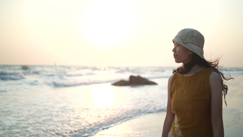 young Asian woman having relax and freedom time, walking enjoy on the tropical sea beach, happy travel in holiday, summer vacation lifestyle concept, ocean scene with sunset, slow-motion shot
