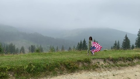 happy little girl is running and jumping carefree with open arms in cloudy mountains. Holding USA flag. selective focus. noise in the shadows.