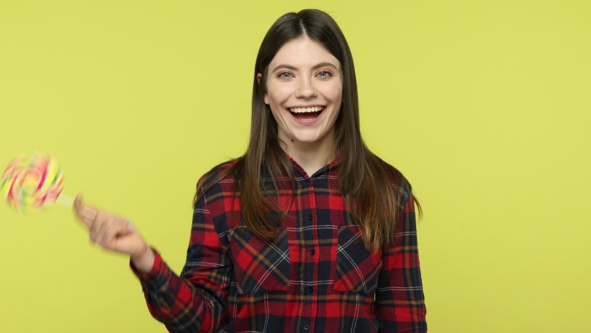 Playful cheerful brunette girl in checkered shirt covering one eye with big colored lollipop and licking it, having fun, enjoying sweets. Indoor studio shot isolated on yellow background