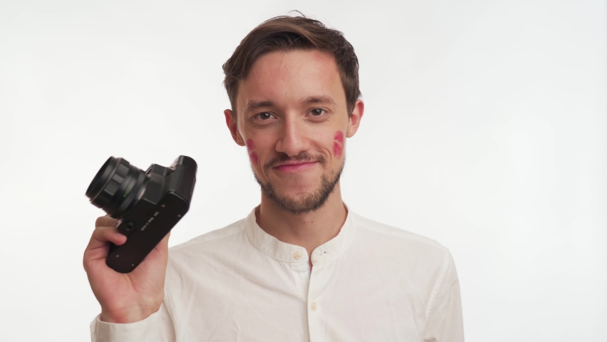 Caucasian bearded brunette man wear shirt smile, hold retro photo camera, take a shot isolated on white wall background close up. Photographer guy kissed by girlfriend, red lipstick kiss marks on face