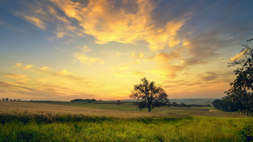 Time Lapse of the rising sun over a tree in an open rural landscape, with clouds in the blue sky beautifully playing with the rays of light
