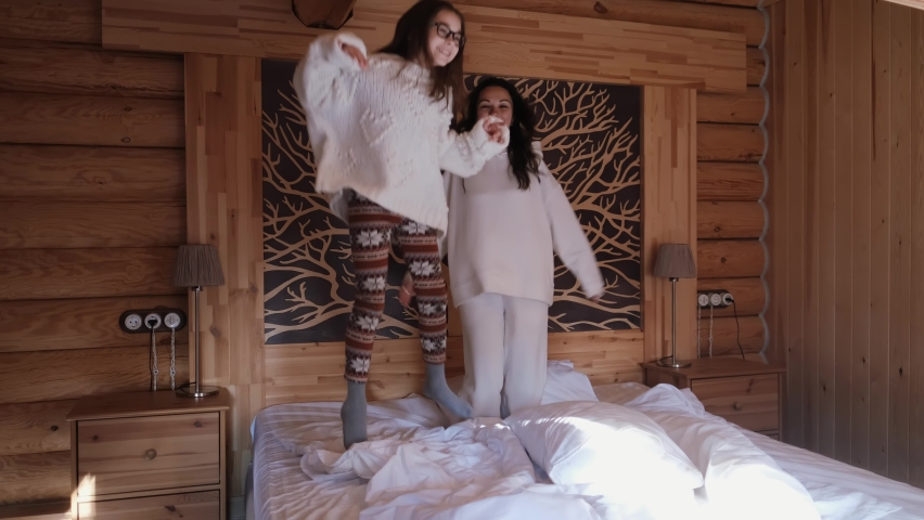 Cute teenage girl and her mom are jumping on the bed and having fun in a cozy country wooden house