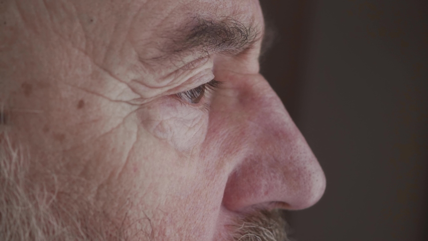 Close up senior man looking to outside from window. Lonely old man looking into distance, waiting for relatives at retirement home. An elderly thoughtful man is looking out a window of his house. Royalty-Free Stock Footage #1067798456