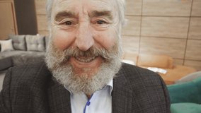 60-something Caucasian bearded man with cropped grey hair sitting on couch at home, holding invisible smartphone in outstretched hand, waving and chatting enthusiastically on video call