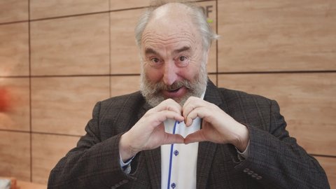 Smiling old bearded man showing hands sign heart shape looking at camera.