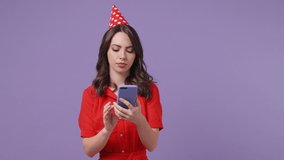 Funny young woman in red dress birthday hat posing isolated on violet background studio. Celebrating holiday party concept. Talking on mobile cell phone making video call greeting with hand thanking