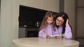 A brunette with black-rimmed glasses sits at the kitchen table with a tablet in her hands, next to her is a little daughter, 6 years old, with long hair.
