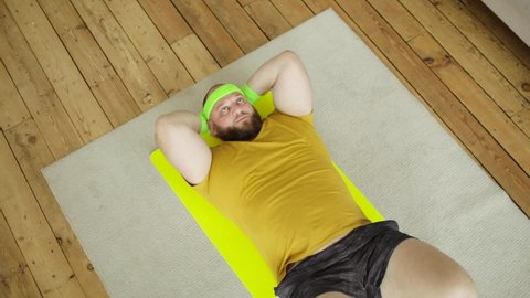 Fat tired man newcomer in sport wearing in yellow sportswear is doing abs exercise at home lying on mat, top view. Joke, mem, humor, parody. Workout, training, fitness, loosing weight concept.