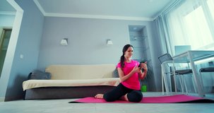 Young attractive fitness woman blogger recording video with a smartphone doing sport exercises or practice yoga on a mat in living room or kitchen at home.