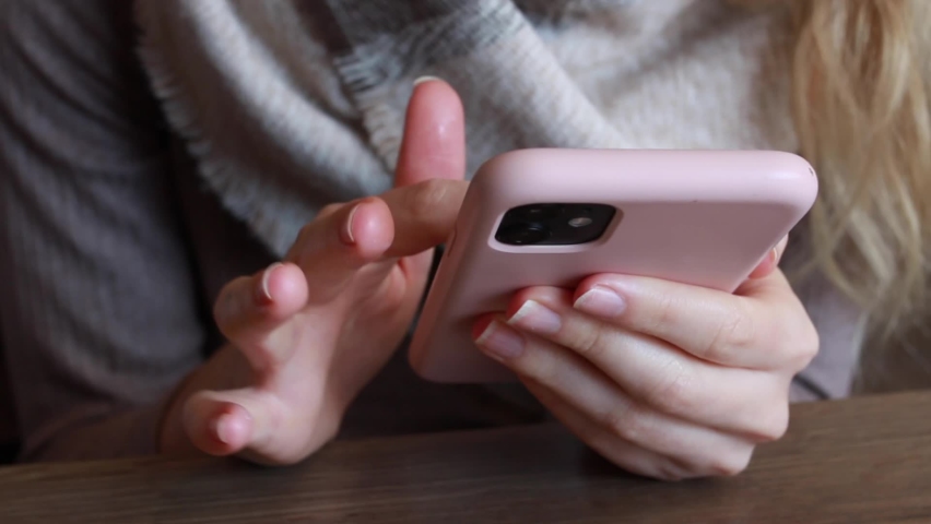 Woman with a finger tattoo and blonde hair scrolling on her pink smartphone rapidly in a cafe, February 2021.