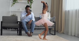Young Afro American parents watch their beloved daughter dance, loving father makes video photo on smartphone camera of mobile phone while little girl princess ballerina dancing whirling in new dress