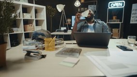 African businessman enjoying smell of cash in modern coworking. Cheerful afro male person kissing bills at dark workplace. Successful african american guy holding pile of money in hipster office.