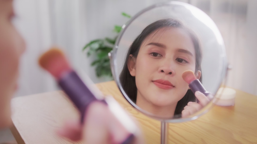 Beautiful attractive young Asian woman doing makeup while looking at mirror putting powder foundation on face using brush, beauty vlogger happy smiling cheerful at home living domestic lifestyle