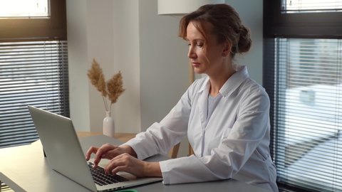Serious young woman doctor physician wearing white coat working typing on laptop computer looking on display screen, sitting at desk in office room at hospital near window. Shooting in slow motion.