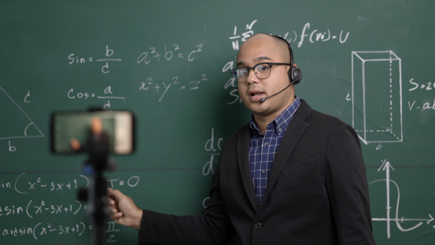 Indian young teacher man wearing headset teaching online video conference live stream by smartphone. Asian teacher teaching mathematics class webinar online for students learning. Royalty-Free Stock Footage #1067843168